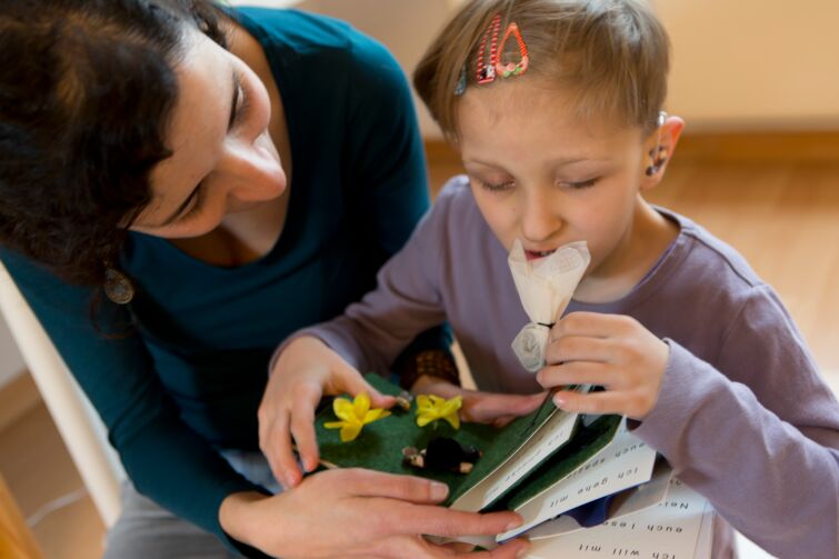 Ein Mädchen im Vorschul-Alter riecht an einem Duftsäckchen, das zu einem Tastbuch für blinde und sehbehinderte Kinder gehört. Sie trägt ein Hörgerät.