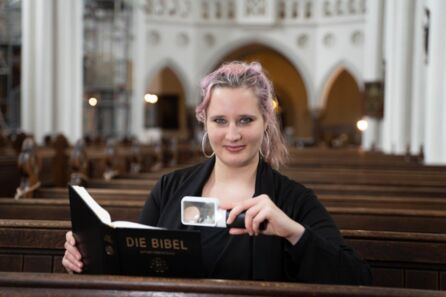 Eine Frau Mitte 20 sitzt in einer Kirchenbank und lächelt in die Kamera, in einer Hand eine Leuchtlupe, in der anderen eine Bibel. Sie hat langes, dunkelblondes Haar mit blonden und rötlichen Strähnchen und trägt große Ohrringe. Bildnachweis "DBSV/Friese"