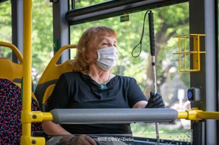Eine Seniorin mit Langstock sitzt in einem Bus. Sie hat rotblonde Haare, trägt eine weiße Mund-Nasen-Maske, ein schwarzes T-Shirt und einen schwarzen Handschuh an der linken Hand, mit der sie den Langstock hält.
