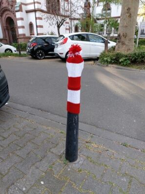 Poller mit rot-weißer Pollermütze, dahinter eine Straße mit parkenden Autos