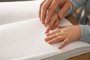 Nahaufnahme: zwei Kinderhände liegen auf einer Seite mit Blindenschrift. Die Hand eines Erwachsenen führt die rechte Kinderhand, deren Zeigefinger ausgestreckt ist. Die linke Kinderhand liegt daneben auf dem Papier.