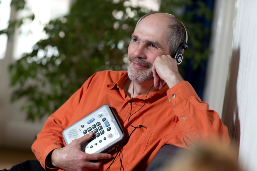 Ein blinder Mann mit Kopfhörern sitzt auf einem Sofa und hält einen DAISY-Player in der Hand