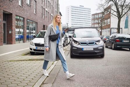 Farbfoto: Eine junge blonde Frau mit grauem Mantel, Jeans und einem Smartphone in der Hand betritt eine Straße, auf der sich ihr ein dunkles Auto nähert. Ihr Blick ist dem Auto abgewandt.