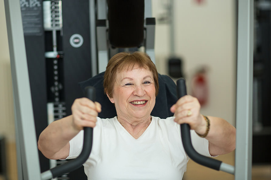 Eine ältere Dame sitzt in einem Fitnessstudio an einem Fitnessgerät. Sie blickt in Richtung des Betrachters und lächelt freundlich.
